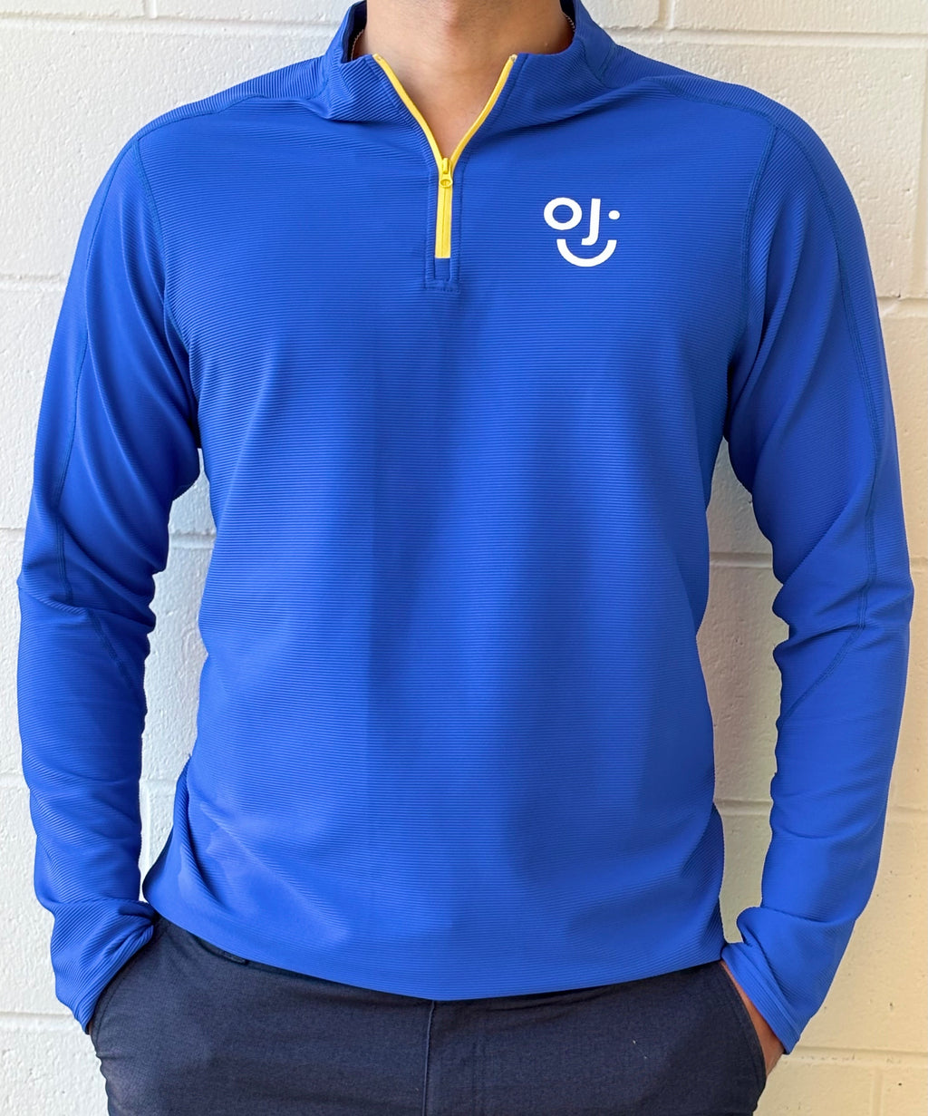 Blue long-sleeve quarter zip with OJI logo on a white brick wall background